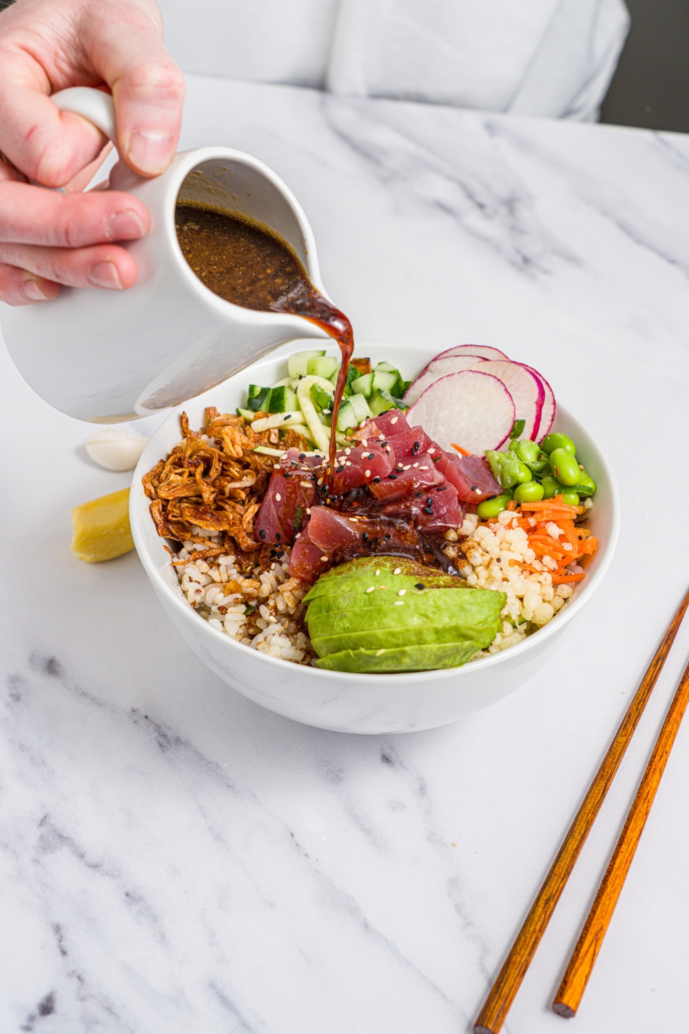 A creamer of poke sauce being poured over a tuna poke bowl. The bowl is served with avocado, rice, crispy onions, radishes, edamame, cucumber, and sesame seeds. The bowl is on a marble counter with a pair of chopsticks.