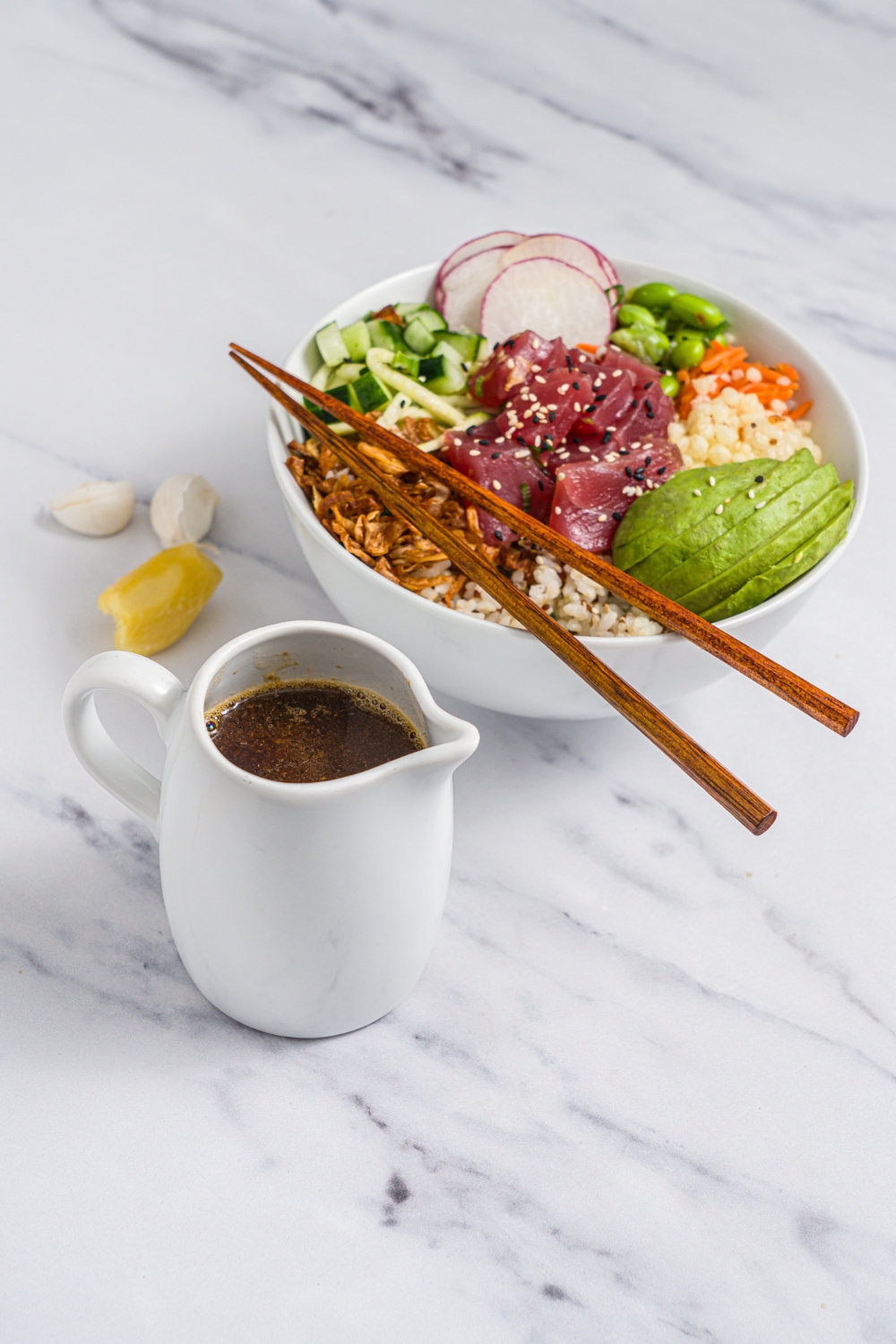 A creamer of poke sauce on a marble counter with a large tuna poke bowl. The bowl is served with rice, avocado, cucumber, radishes, edamame, crispy onions, sesame seeds, and chopsticks.