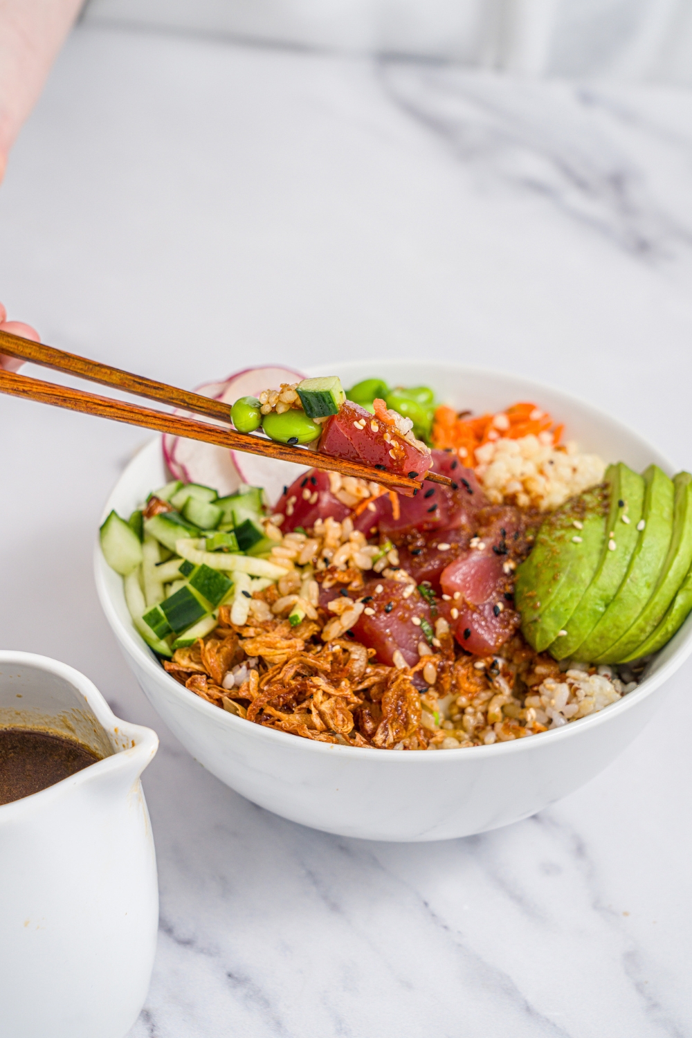 A tuna poke bowl in a white bowl with rice, avocado, edamame, crispy onions, cucumber radishes, and sesame seeds. A pair of chopsticks in taking a bite from the bowl. The bowl is on a marble counter with a creamer of poke sauce.