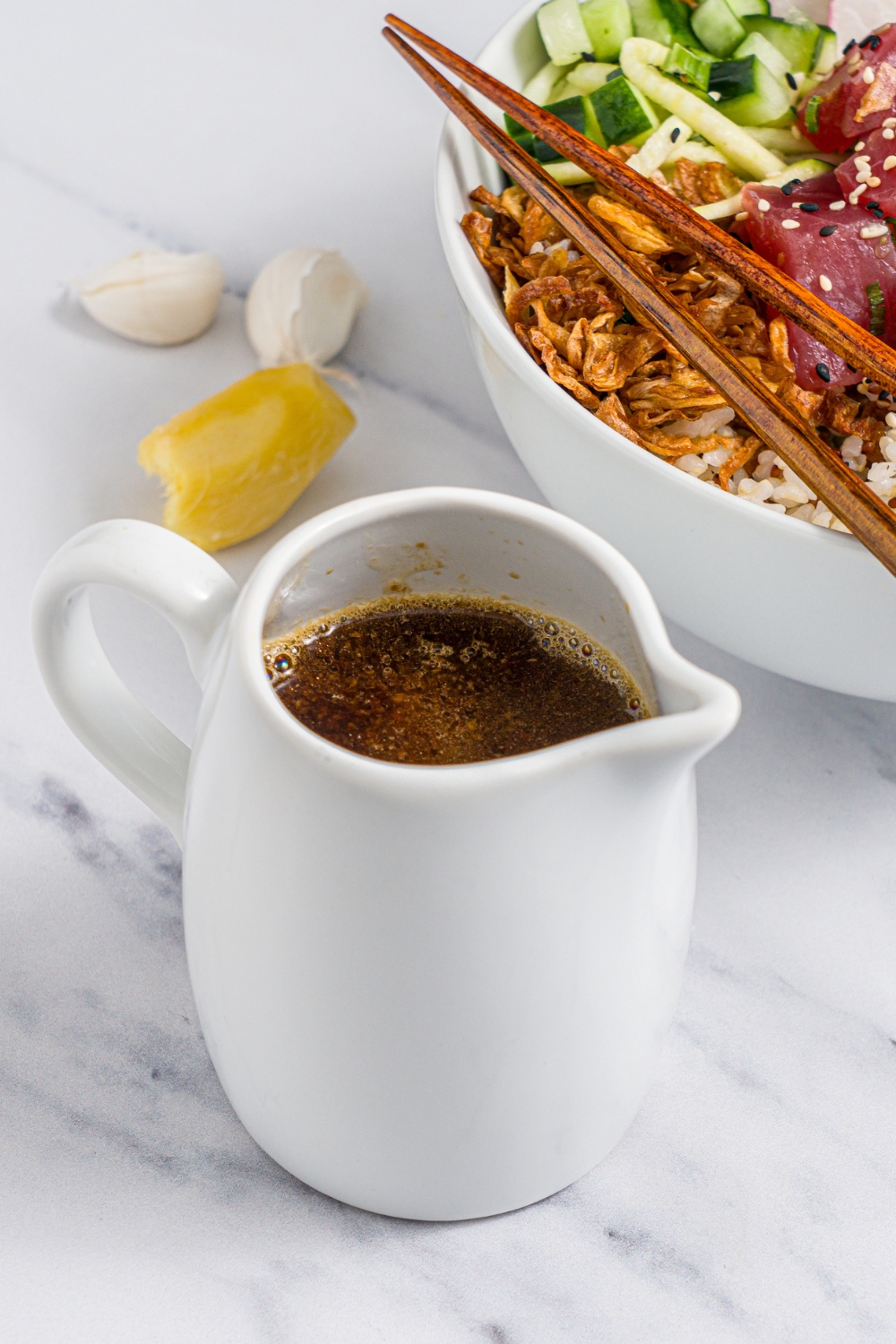 A small creamer filled with poke sauce. The creamer is on a white counter with a tuna poke bowl, garlic, and ginger.