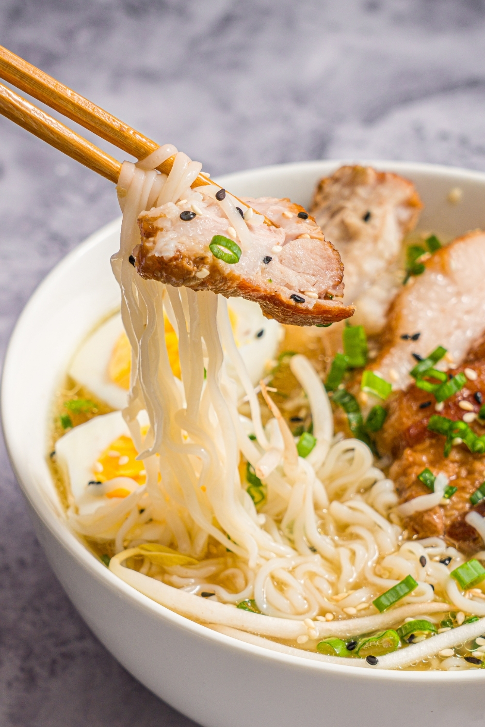 A large white bowl with pork belly miso ramen topped with bean sprouts, sliced green onion, sliced hard boiled egg, and sesame seeds. A pair of chopsticks is taking a bite of ramen from the bowl. The bowl is on a marble counter.