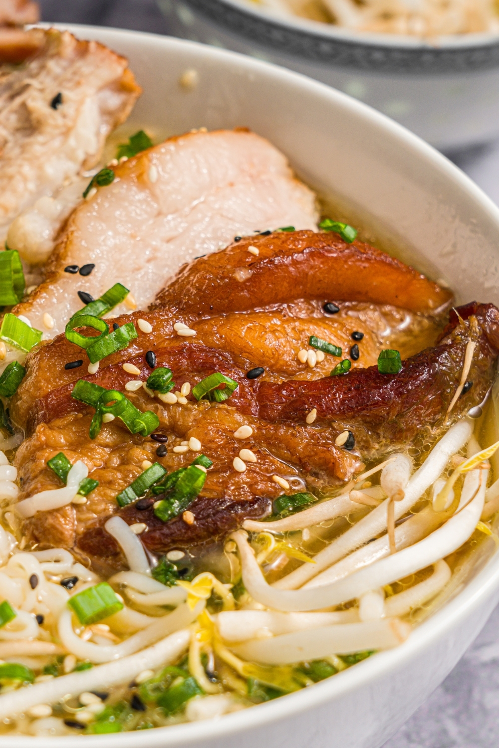 A large white bowl with pork belly miso ramen topped with bean sprouts, green onion, and sesame seeds. The bowl is on a marble counter with small bowls of ingredients.