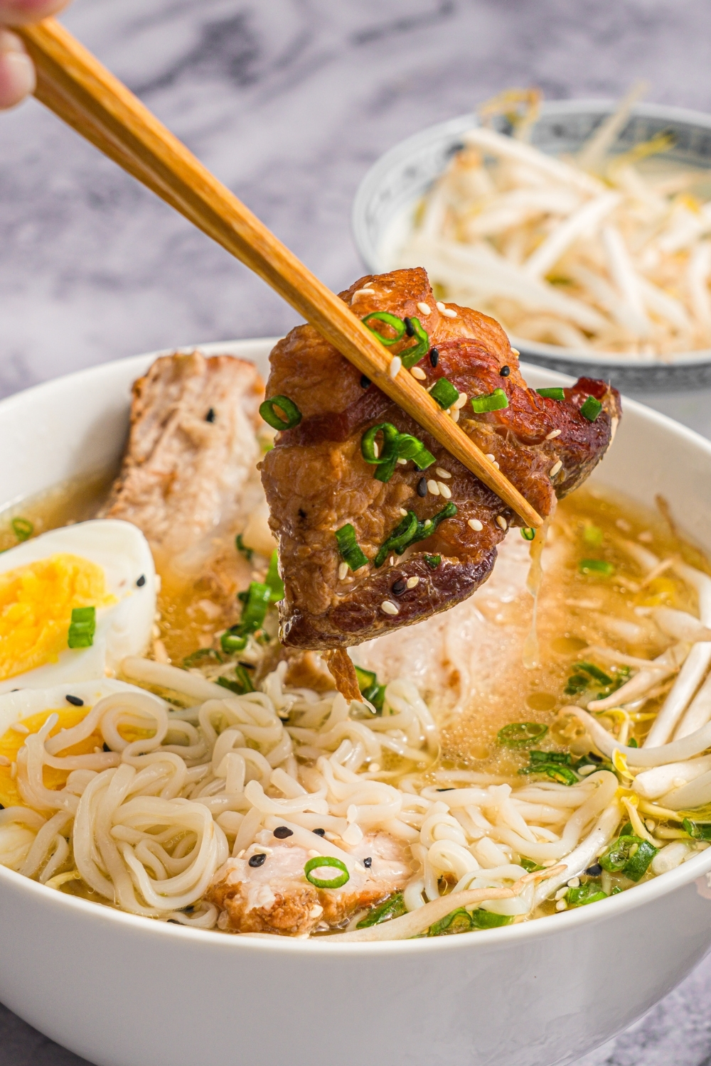 A large white bowl with pork belly miso ramen topped with bean sprouts, green onion, sliced hard boiled egg, and sesame seeds. A pair of chopsticks is holding a piece of pork belly over the bowl. The bowl is on a marble counter with small bowls of ingredients.