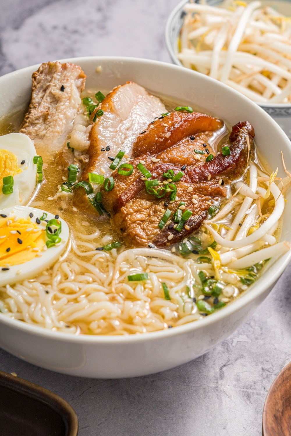 A large white bowl with pork belly miso ramen topped with bean sprouts, green onion, sliced hard boiled egg, and sesame seeds. The bowl is on a marble counter with small bowls of ingredients.