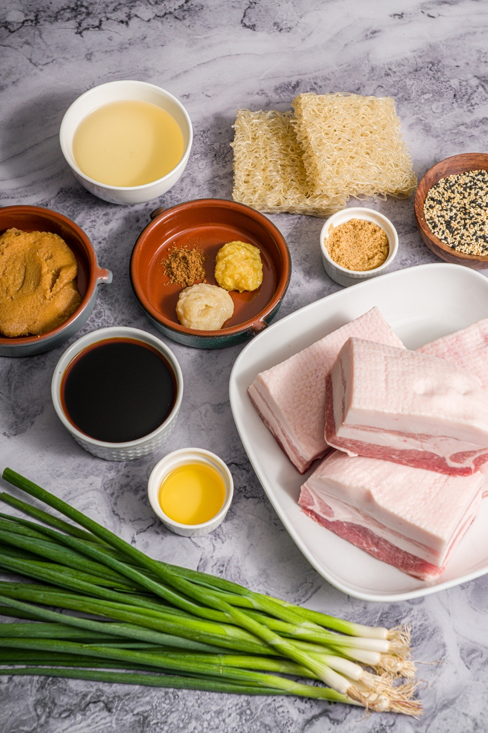 A marble counter with multiple bowls in various sizes containing ingredients to make pork belly ramen including pork belly, scallion, ginger, ramen noodles, garlic paste, ginger paste, miso paste, mirin, soy sauce, brown sugar, chicken broth, sesame oil, and seasonings.