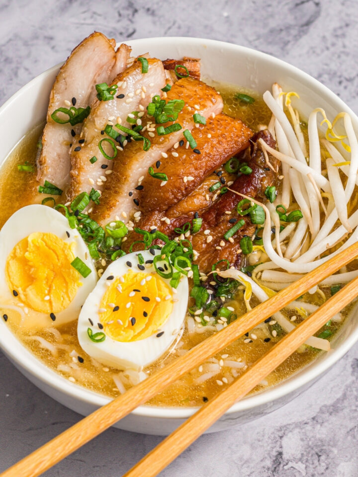 A large white bowl with pork belly miso ramen topped with bean sprouts, green onion, sliced hard boiled egg, and sesame seeds. A pair of chopsticks are resting on the bowl. The bowl is on a marble counter.