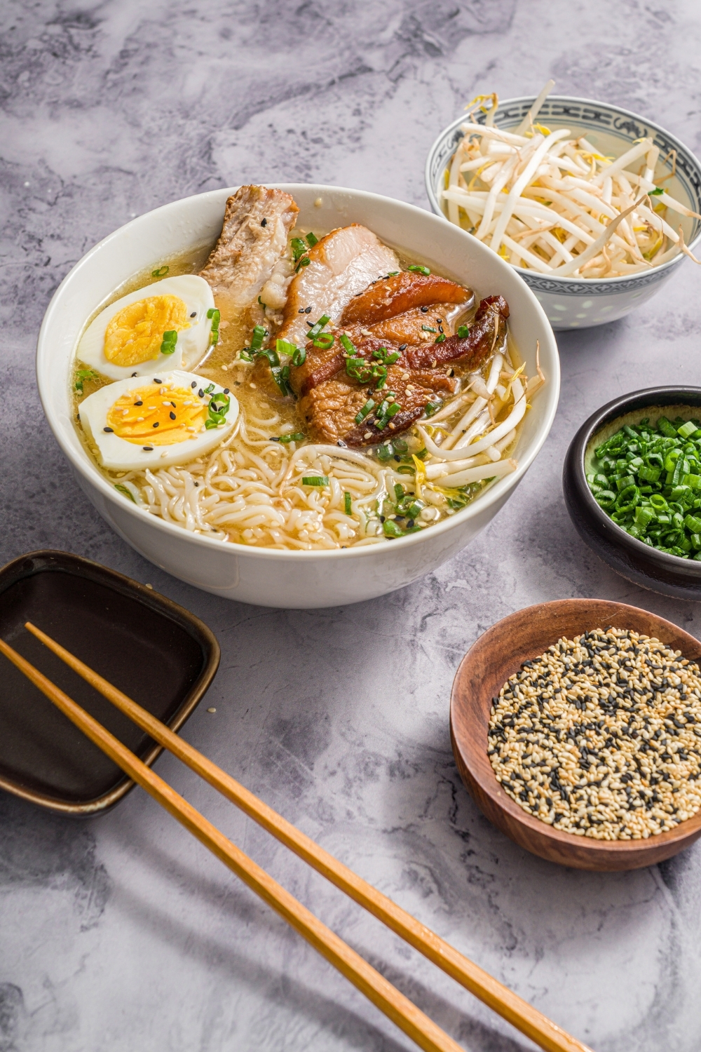 A large white bowl with pork belly miso ramen topped with bean sprouts, green onion, sliced hard boiled egg, and sesame seeds. The bowl is on a marble counter with a pair of chopsticks and small bowls of ingredients.