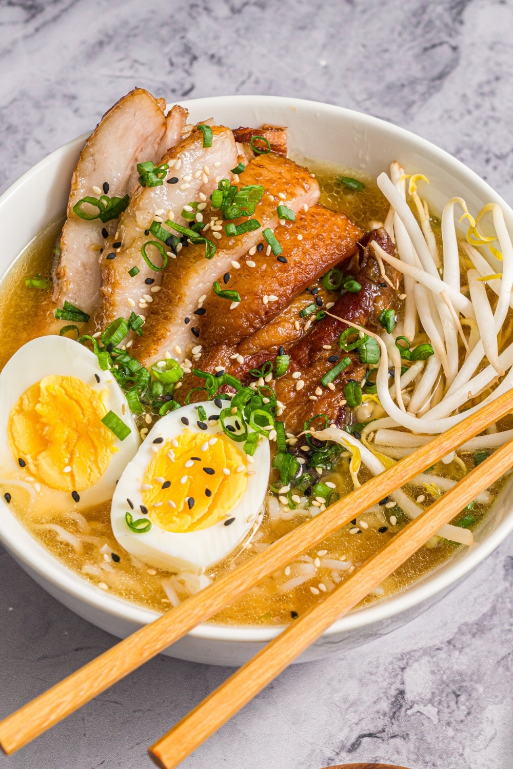 A large white bowl with pork belly miso ramen topped with bean sprouts, green onion, sliced hard boiled egg, and sesame seeds. A pair of chopsticks are resting on the bowl. The bowl is on a marble counter.