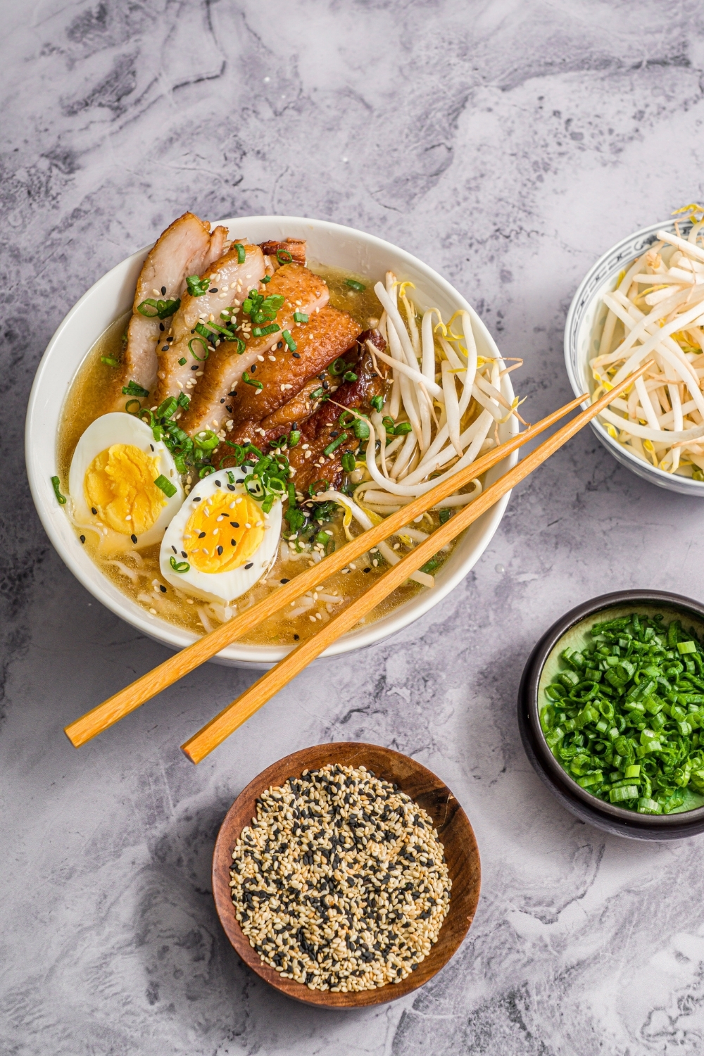 A large white bowl with pork belly miso ramen topped with bean sprouts, green onion, sliced hard boiled egg, and sesame seeds. A pair of chopsticks is resting on the bowl. The bowl is on a marble counter with small bowls of ingredients.