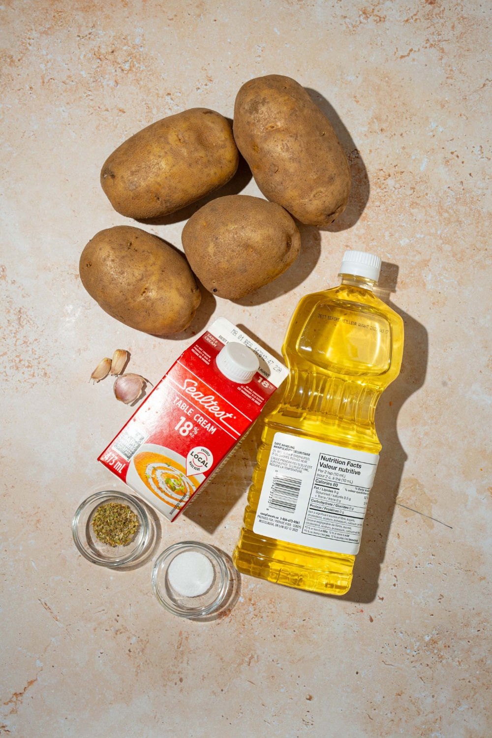 A tan counter with various ingredients to make potato pave including russet potatoes, heavy cream, oil, garlic, and seasonings.