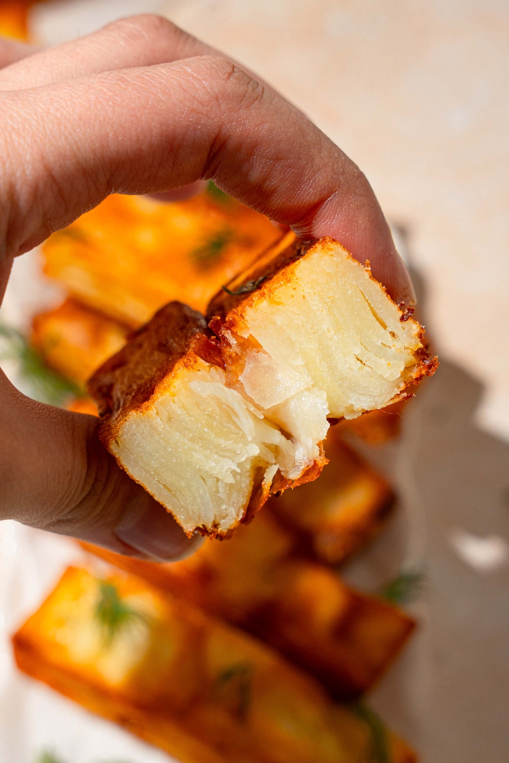A close up of a hand holding a piece of potato pave folded in half. There is a plate with potato pave garnished with fresh thyme blurred in the background.