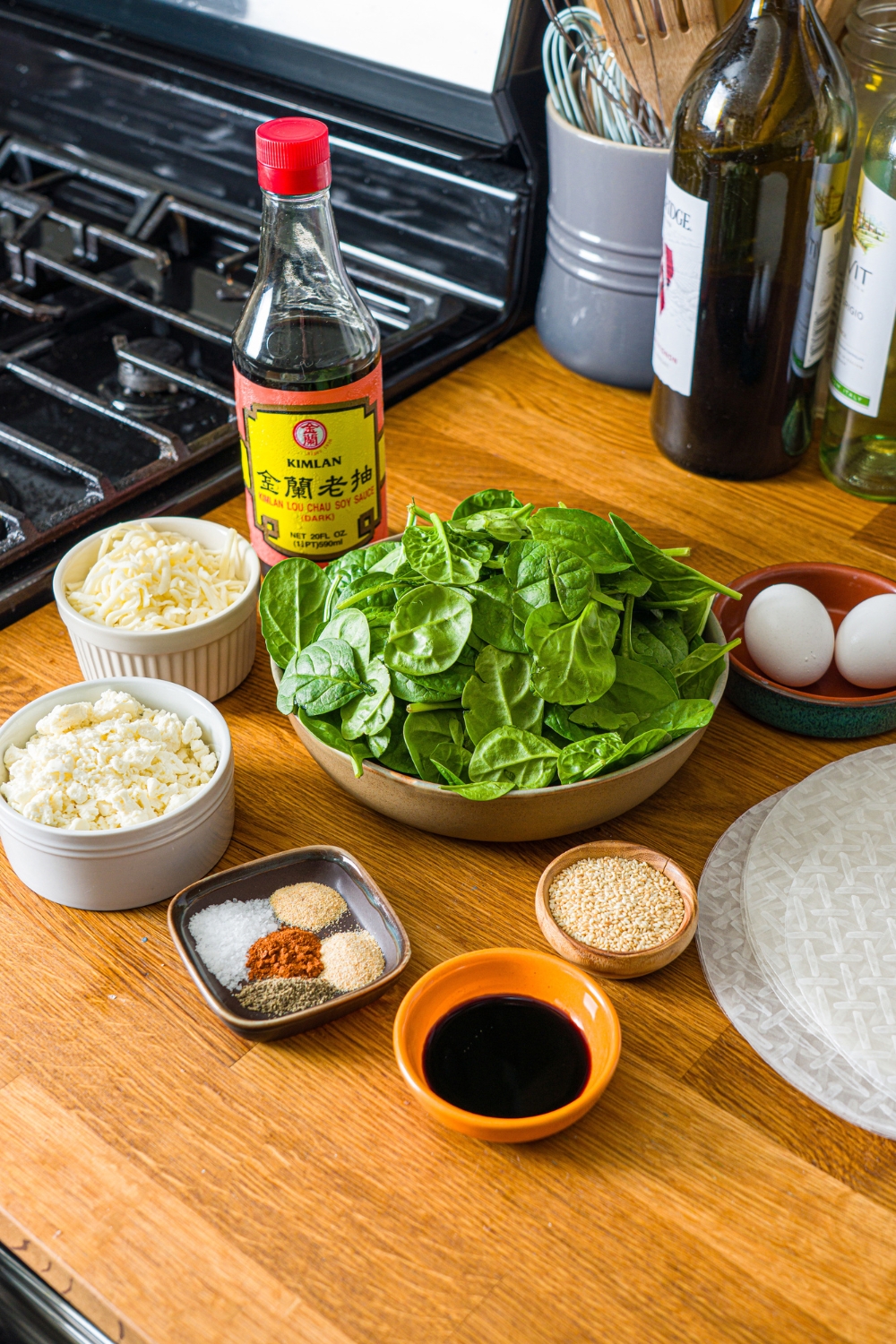 A wooden counter with several bowls in various sizes containing ingredients to make rice paper spanakopita including spinach, rice paper, soy sauce, egg, feta cheese, mozzarella cheese, sesame seeds, and seasonings.