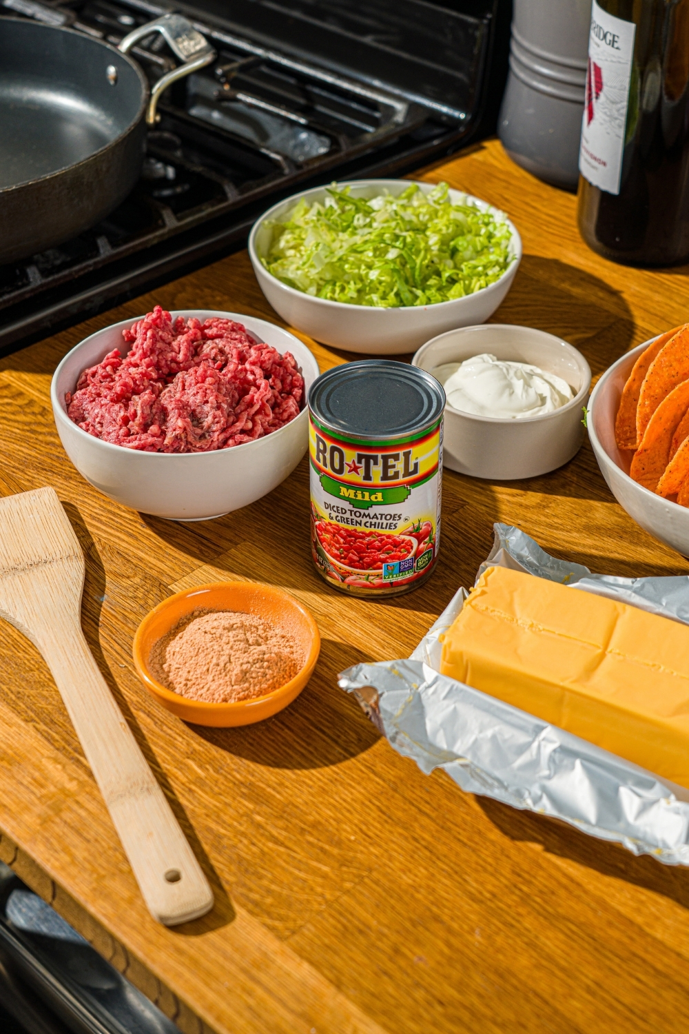 A wooden counter with ingredients to make Rotel tacos including a can of Rotel, velveeta cheese, ground beef, shredded lettuce, taco seasoning, and sour cream.