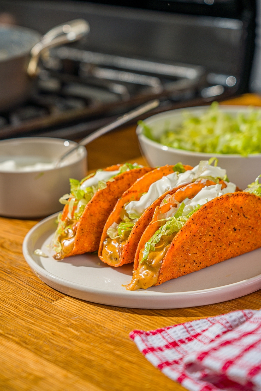 A white plate with three Rotel tacos with a hard shell topped with shredded lettuce and sour cream. The plate is on a wooden counter with a red striped napkin and bowls of toppings.