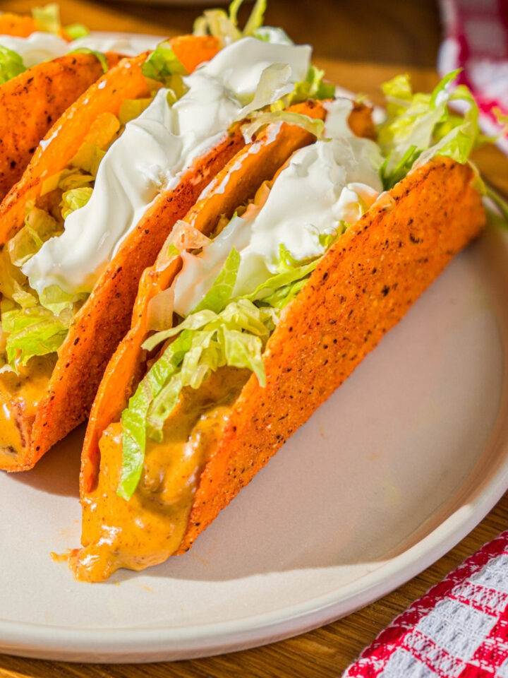 A white plate with three Rotel tacos with a hard shell topped with shredded lettuce and sour cream. The plate is on a wooden counter with a red striped napkin.