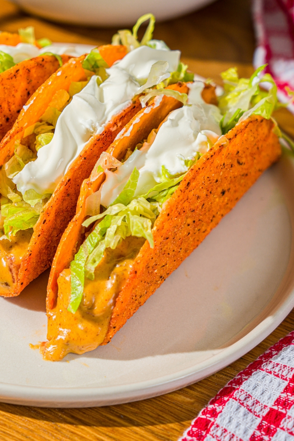 A white plate with three Rotel tacos with a hard shell topped with shredded lettuce and sour cream. The plate is on a wooden counter with a red striped napkin.