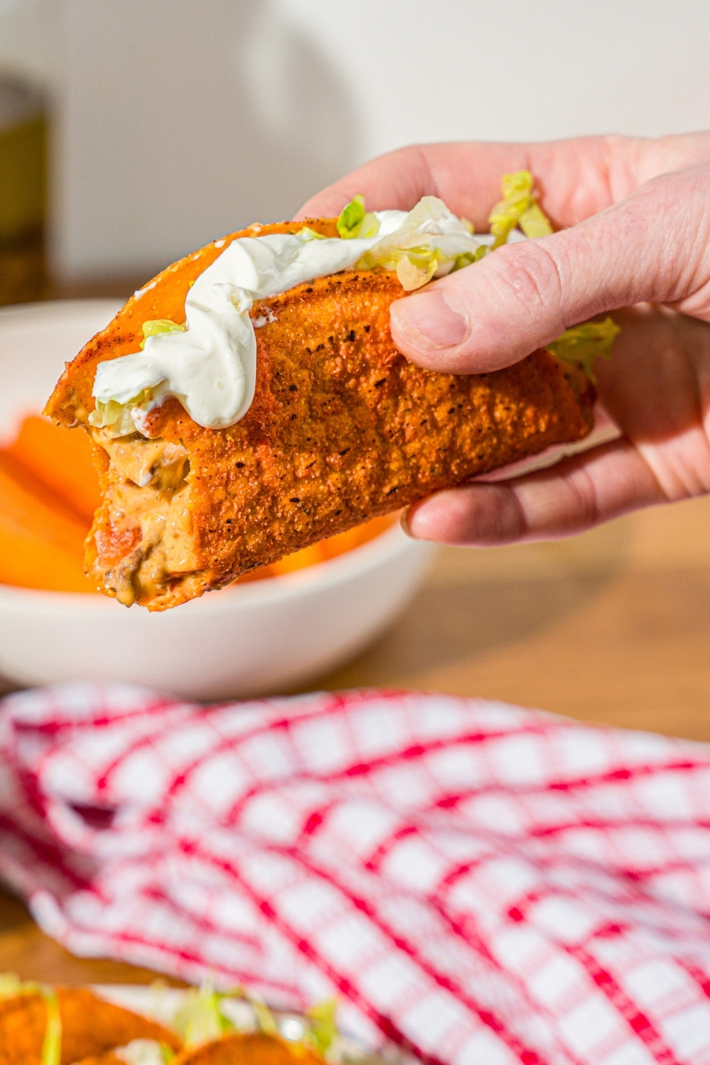 A hand holding a Rotel taco in a hard shell topped with shredded lettuce and sour cream. There is a bite taken from the taco. There is a wooden counter with a red striped napkin in the background.