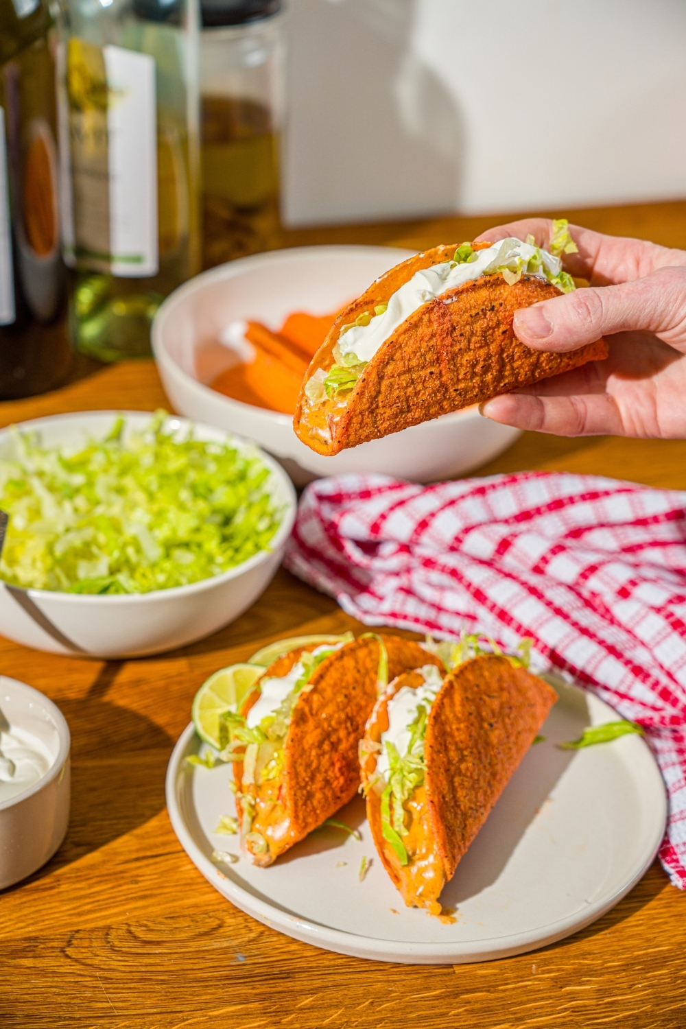 A hand holding a Rotel taco in a hard shell topped with shredded lettuce and sour cream over a plate of tacos. The plate is on a wooden counter with a red striped napkin and bowls of toppings.