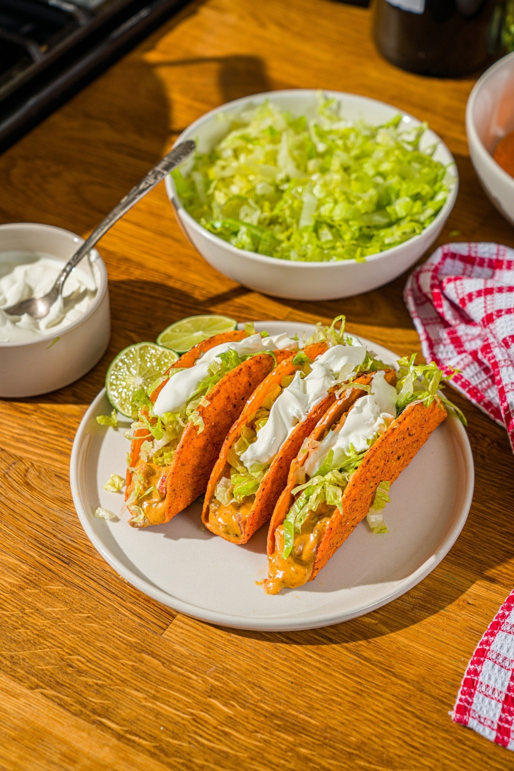 A white plate with three Rotel tacos in a hard shell topped with shredded lettuce and sour cream. The plate is on a wooden counter with a red striped napkin and bowls of toppings.