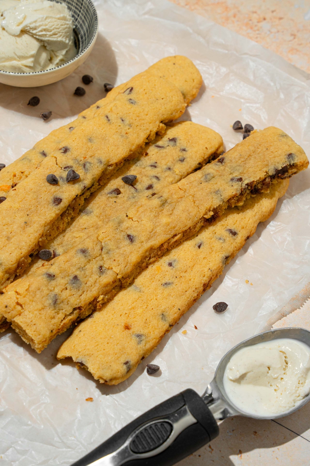 A sheet of parchment paper with several baked chocolate chip cookie fries sprinkled with chocolate chips. There is a bowl of vanilla ice cream and ice cream scoop on the sheet.