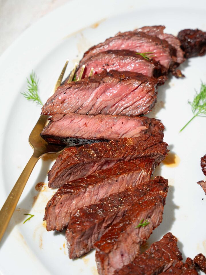 A white plate with sliced marinated skirt steak. There is a fork on the plate. The plate is on a tan counter.