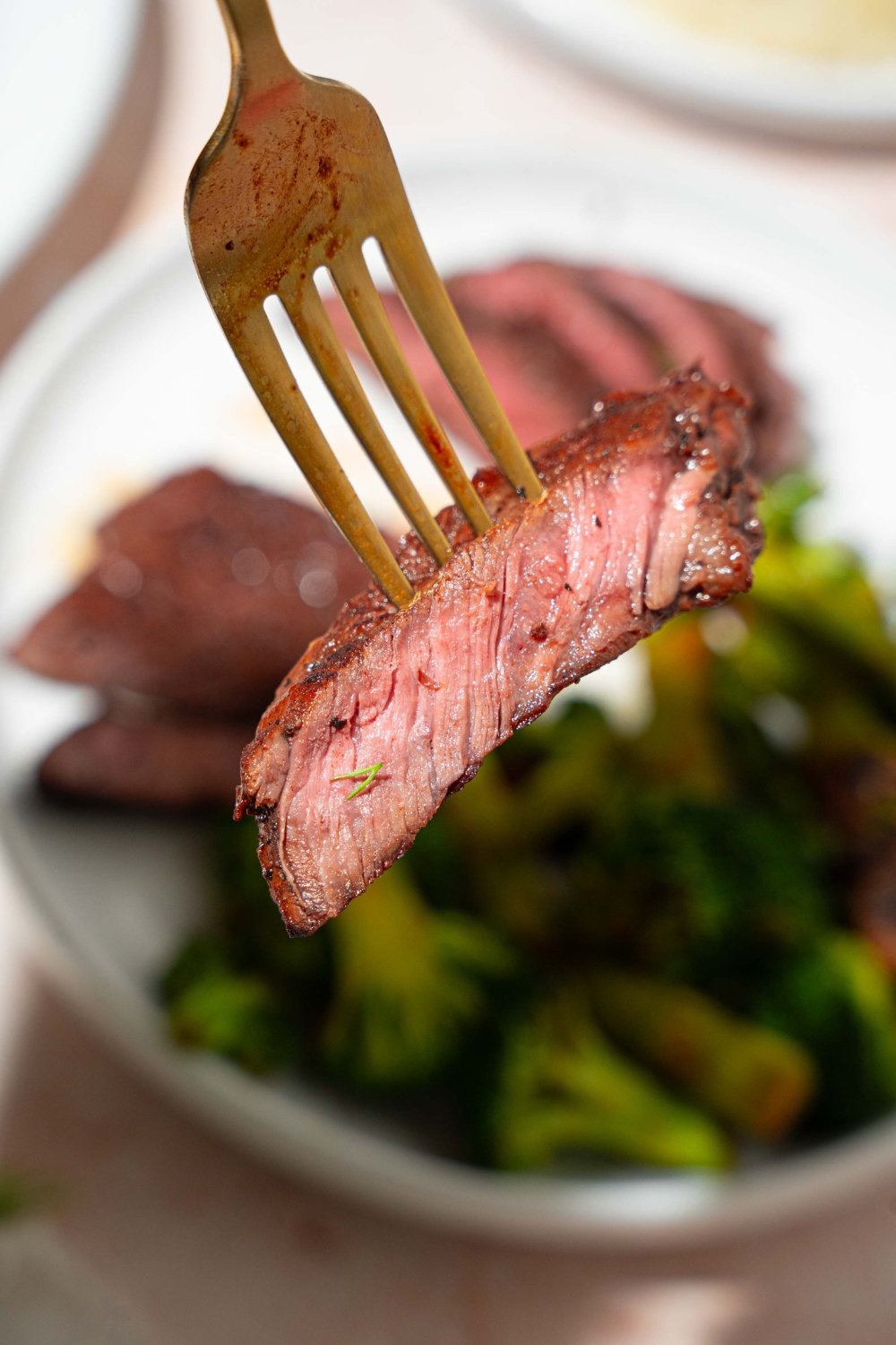 A close up of a fork with a slice of marinated skirt steak. There is a white plate with steak and broccoli blurred in the background.
