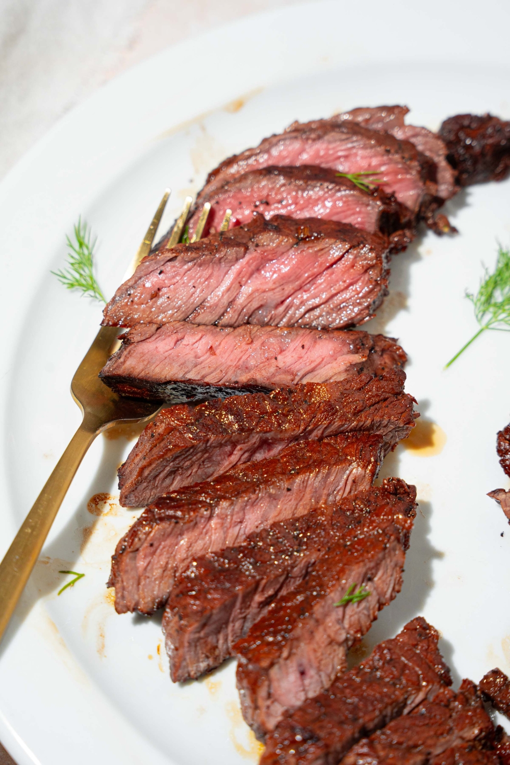 A white plate with sliced marinated skirt steak. There is a fork on the plate. The plate is on a tan counter.