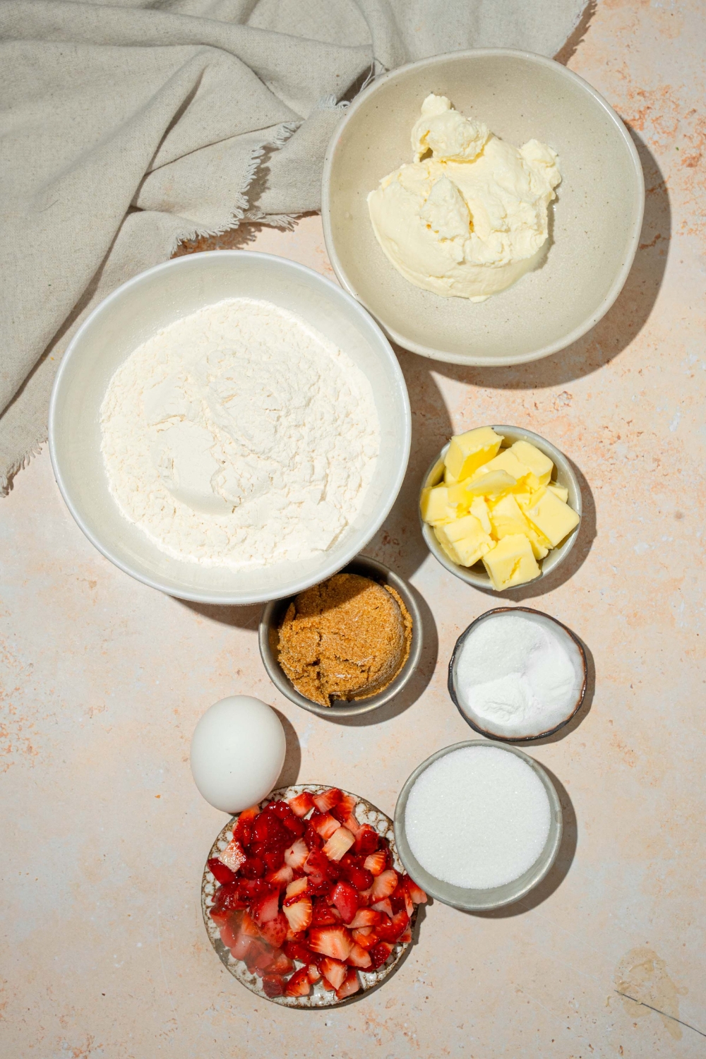 A tan counter with several bowls in various sizes containing ingredients to make strawberry cheesecake cookies including flour, baking powder, baking soda, cream cheese, sugar, brown sugar, egg, butter, and strawberries.