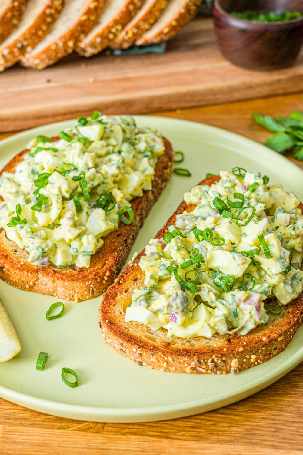 A green plate with two slices of toasted bread topped with pickle egg salad garnished with sliced green onion and served with a pickle spear. The plate is on a wooden counter with a wooden board of sliced bread with garnishes.
