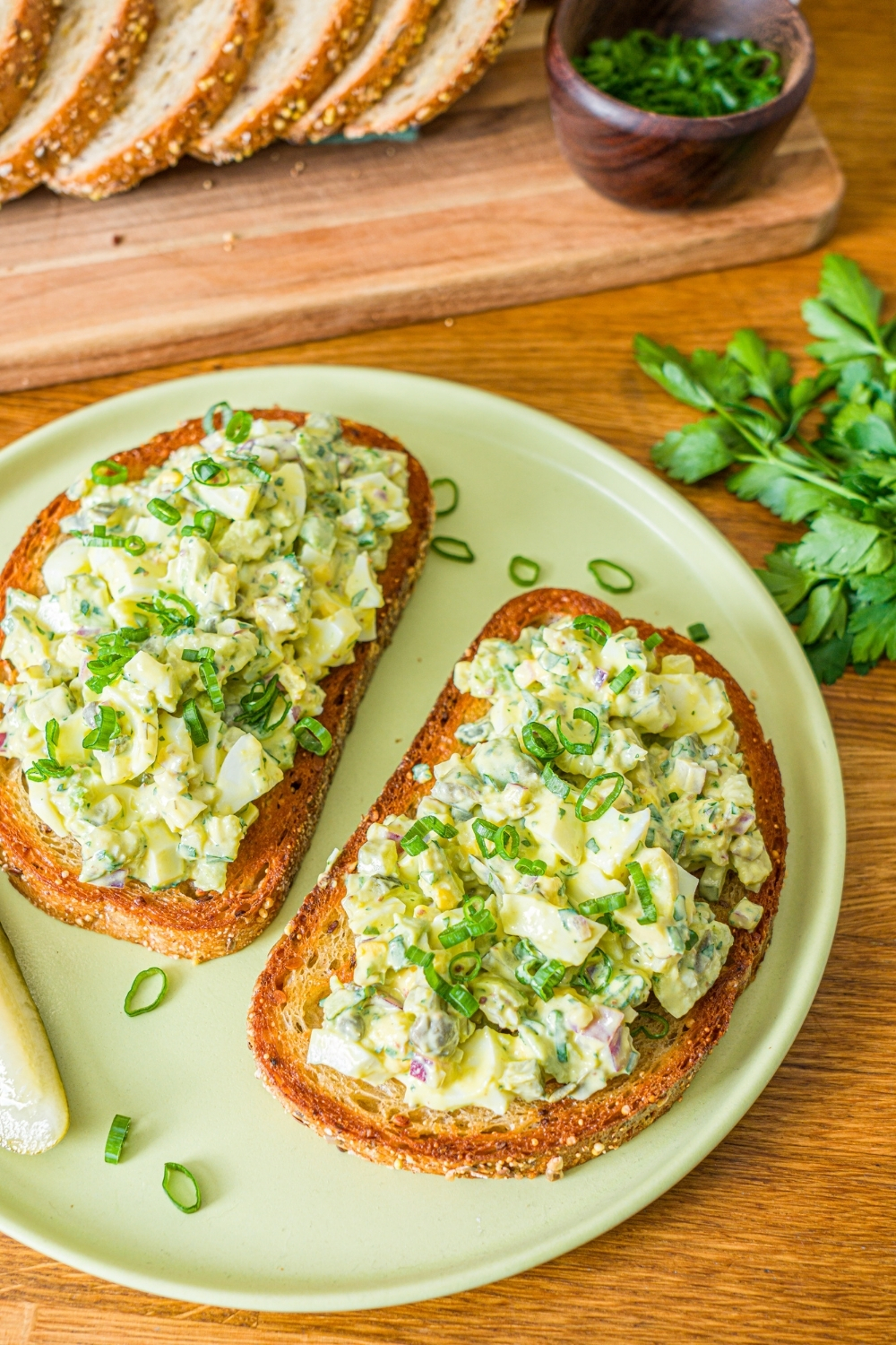 A green plate with two slices of toasted bread topped with pickle egg salad garnished with sliced green onion and served with a pickle spear. The plate is on a wooden counter with a wooden board of sliced bread with garnishes.