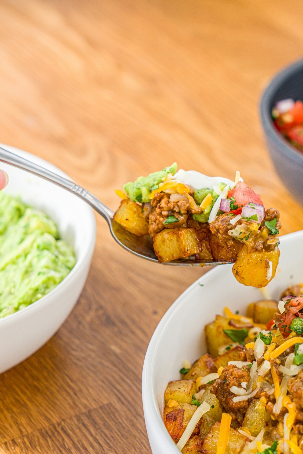 A fork with a bite of loaded potato taco bowl over a white bowl including roasted potatoes, taco meat, shredded cheese, pico de gallo, sour cream, and guacamole. The bowl is garnished with cilantro.