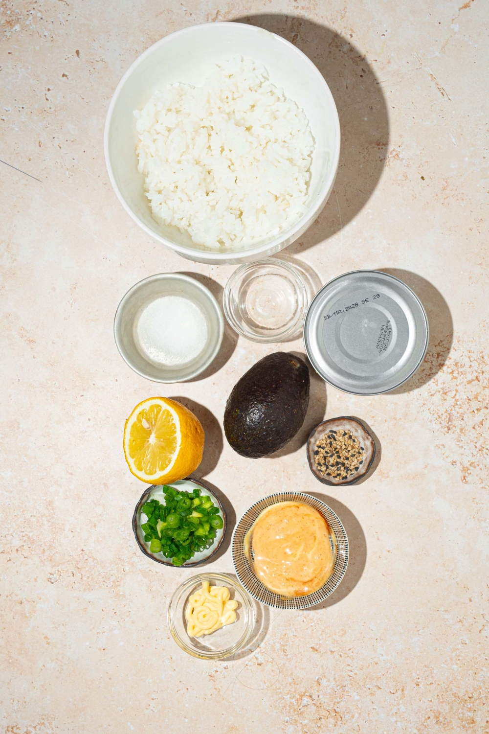 A tan counter with several bowls in various sizes containing ingredients to make tuna rice cups including rice, canned tuna, lemon, mayo, sesame seeds, spicy mayo, avocado, and green onion.