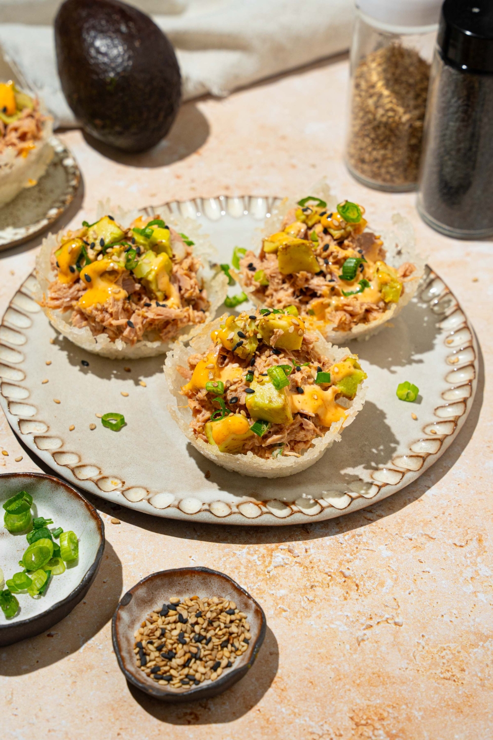 A plate with three tuna rice cups topped with avocado, sliced green onion, sesame seeds, and spicy mayo. The plate is on a tan counter with small plates of garnishes.