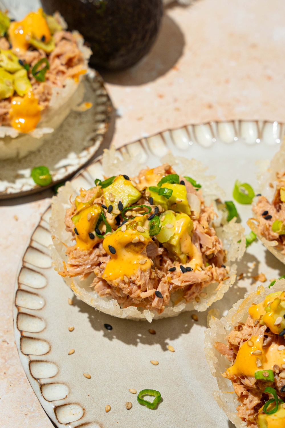 A plate with three tuna rice cups topped with avocado, sliced green onion, sesame seeds, and spicy mayo. The plate is on a tan counter.