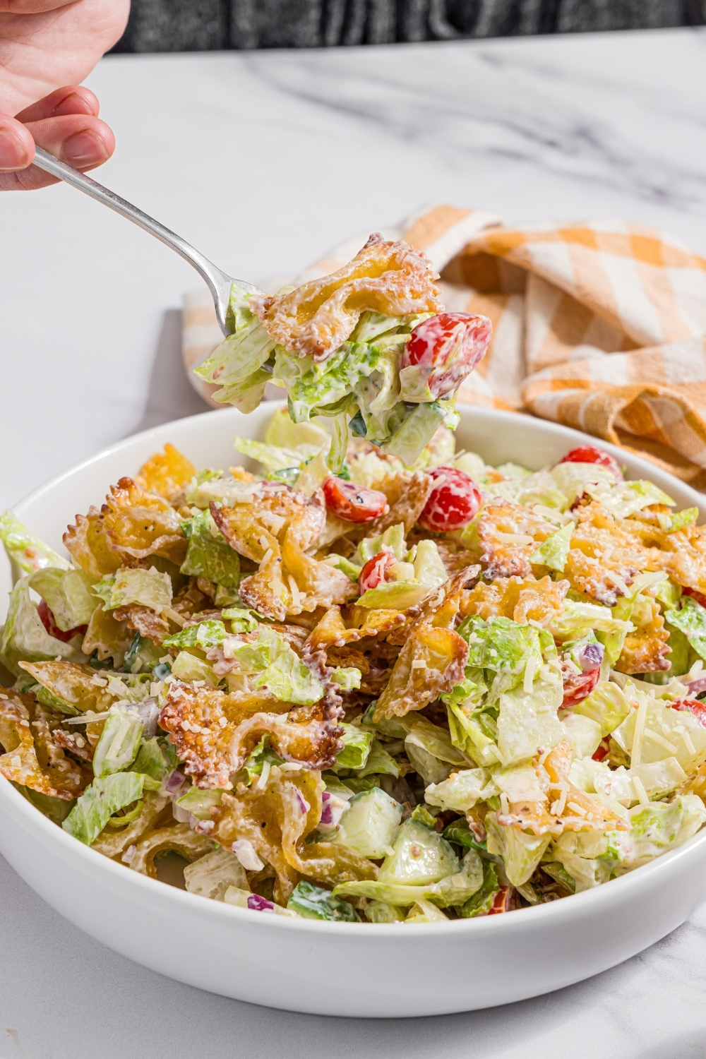 A white bowl with crispy pasta salad tossed with shredded lettuce, tomatoes, cucumber, and onion in a yogurt dressing. There is a fork with a bite of pasta salad over the bowl. The bowl is on a marble counter with a yellow checkered napkin.