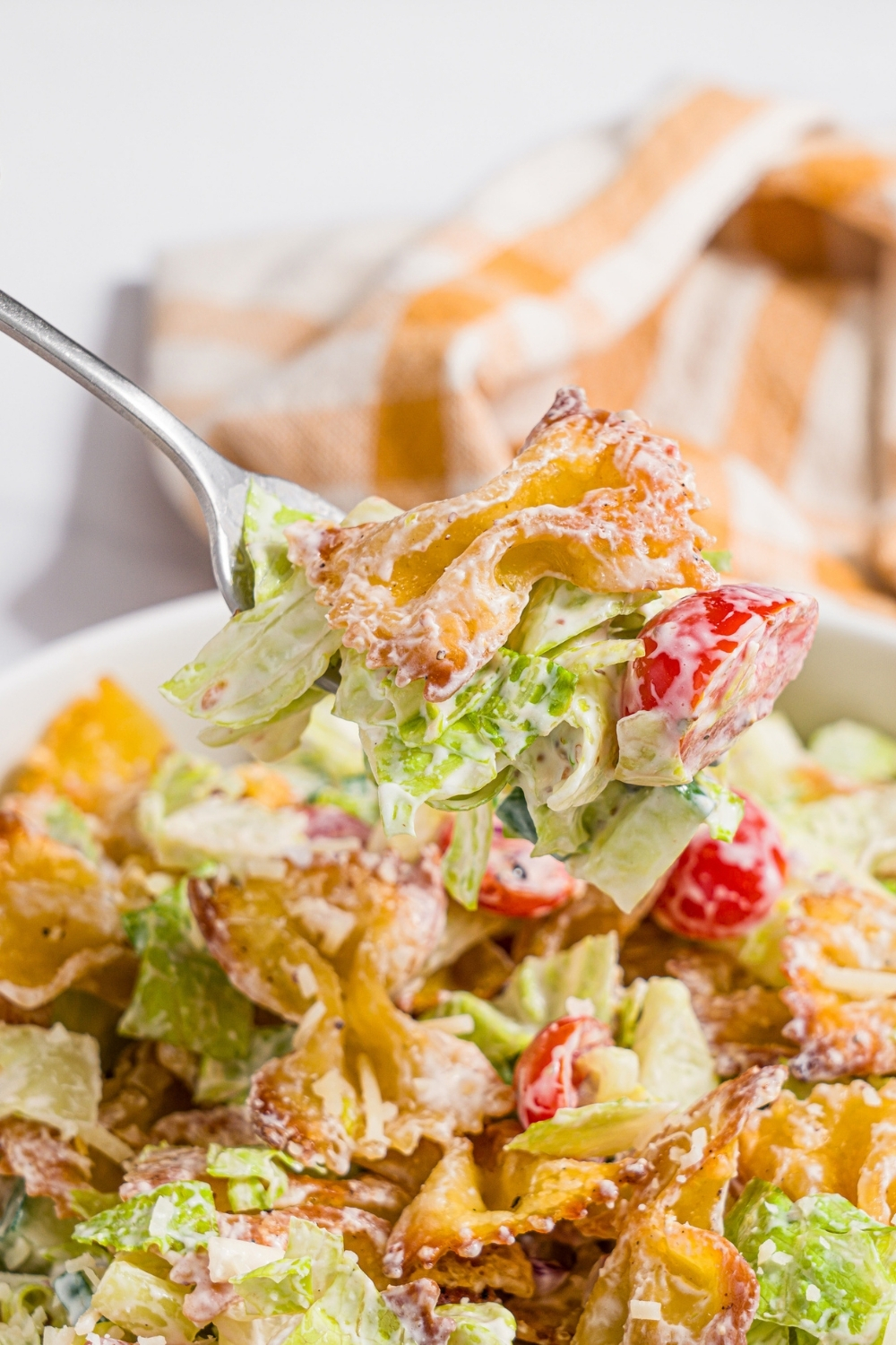 A fork with a bite of crispy pasta salad. There is a white bowl with crispy pasta salad tossed with shredded lettuce, tomatoes, cucumber, and onion in a yogurt dressing. The bowl is on a marble counter with a yellow checkered napkin.