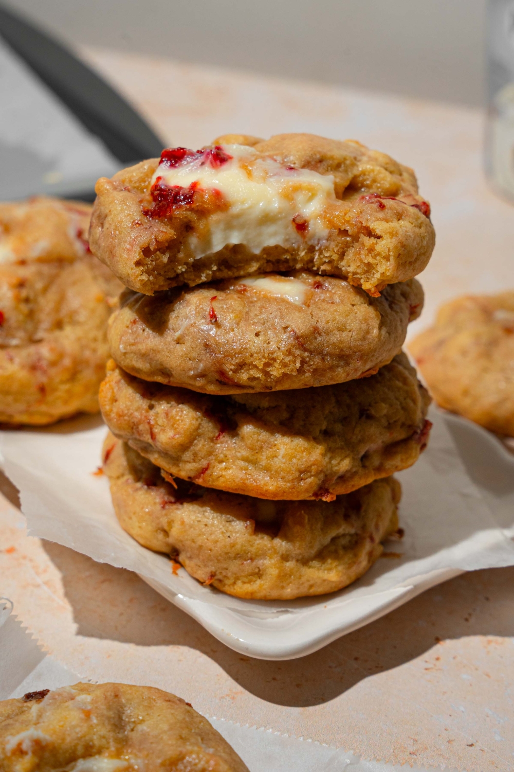 A white plate lined with parchment paper with several strawberry cheesecake cookies stacked on one another. The top cookie has a bite taken from it. The plate is on a tan counter with additional cookies.