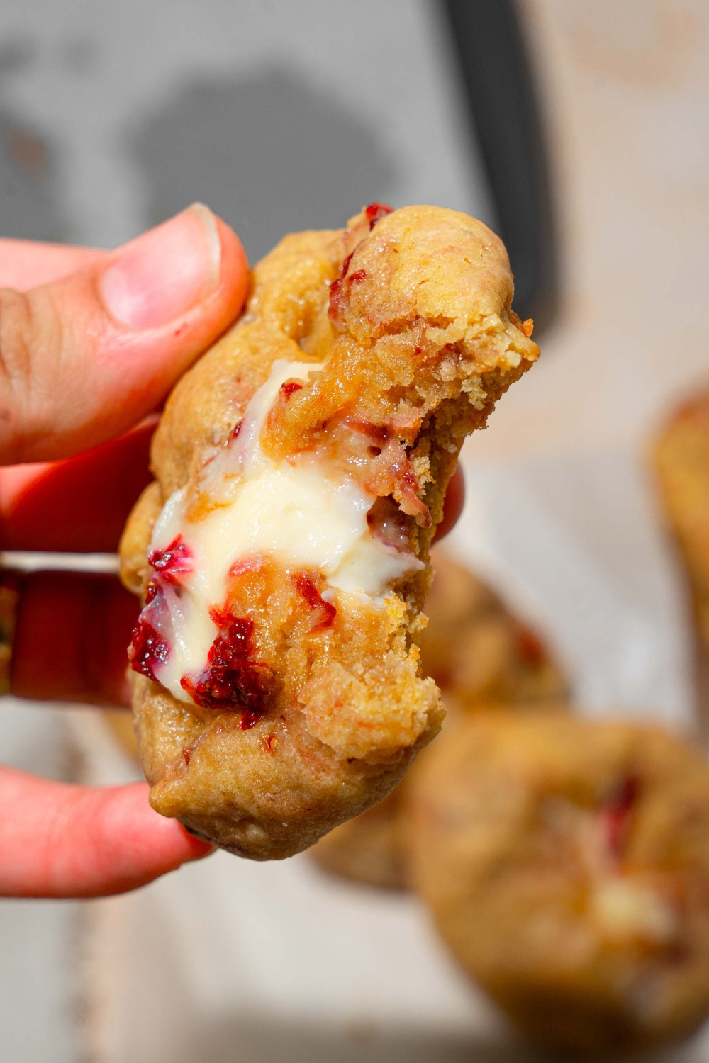 A close up of a strawberry cheesecake cookie with a bite taken from it. There is a white plate with cookies blurred in the background.