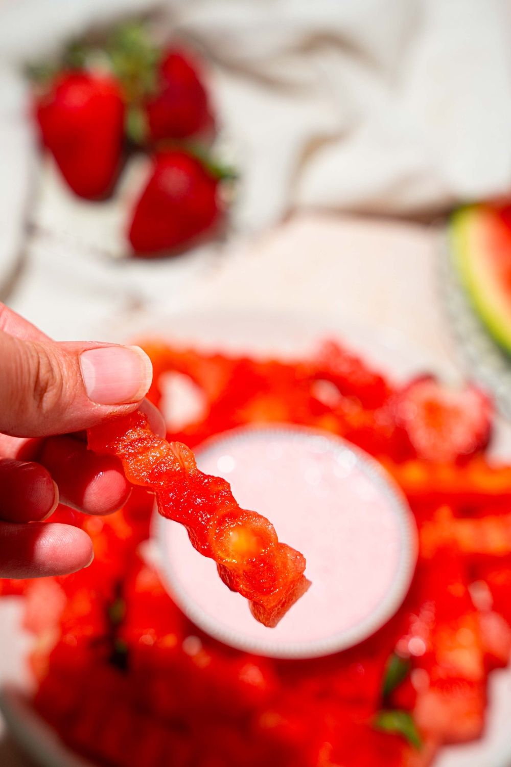 A close up of a hand holding a watermelon fry. There is a plate of watermelon fries with a bowl of strawberry dip blurred in the background.
