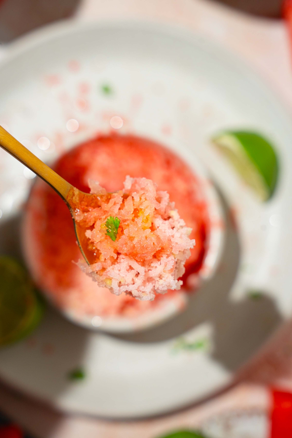 A close up of a spoon with a bite of watermelon shaved ice. There is a bowl of shaved ice on a white plate blurred in the background.