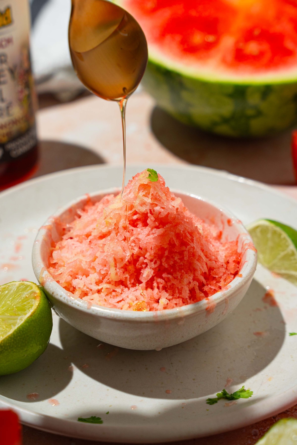 A small white bowl with watermelon shaved ice being drizzled with honey. The bowl is on a white plate with lime. The plate is on a tan counter with watermelon.