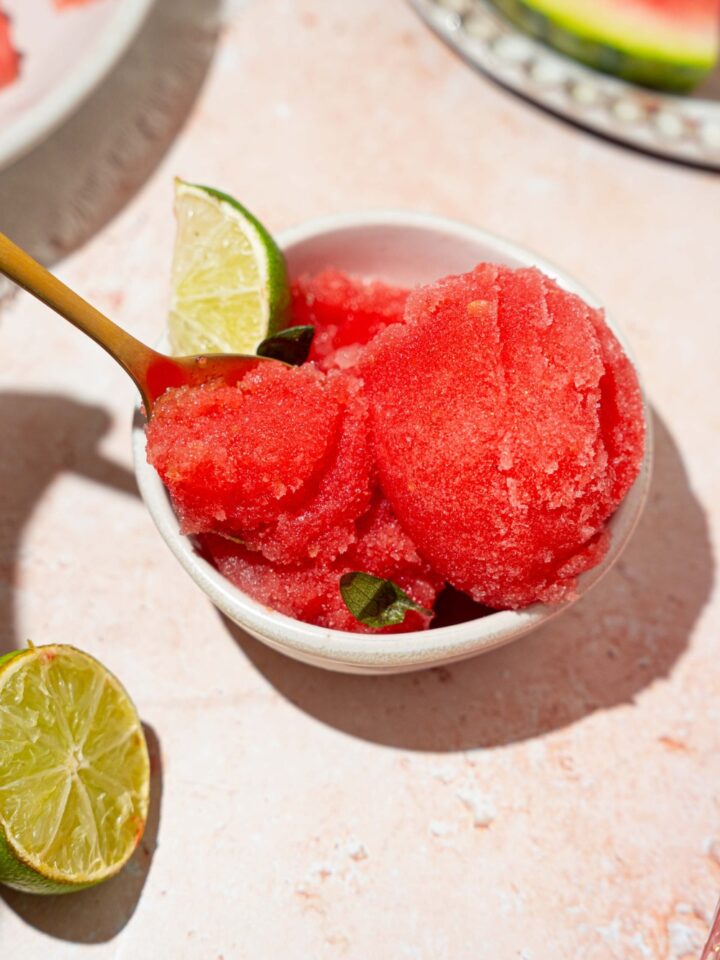 A white bowl with watermelon sorbet served with a lime wedge. There is a spoon in the bowl. The bowl is on a tan counter with lime and a plate of watermelon.