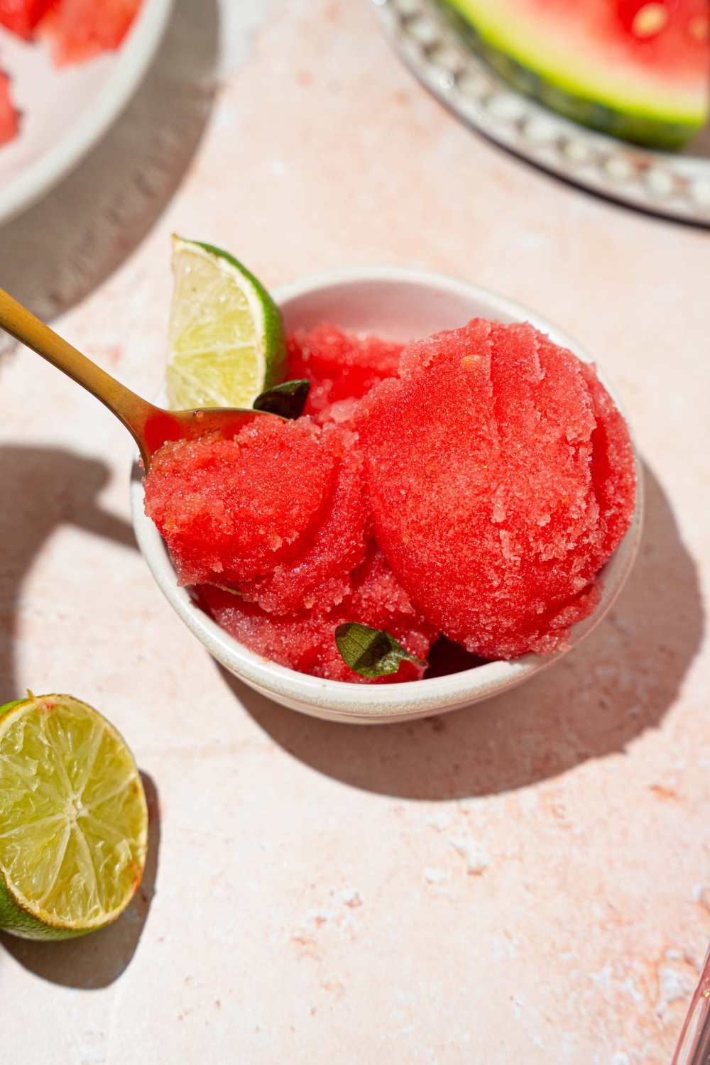 A white bowl with watermelon sorbet served with a lime wedge. There is a spoon in the bowl. The bowl is on a tan counter with lime and a plate of watermelon.