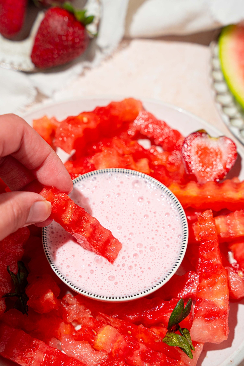 A hand dipping a watermelon fry into a bowl of strawberry yogurt dip. The bowl is on a white plate with watermelon fries and sliced strawberries. The plate is on a tan counter.