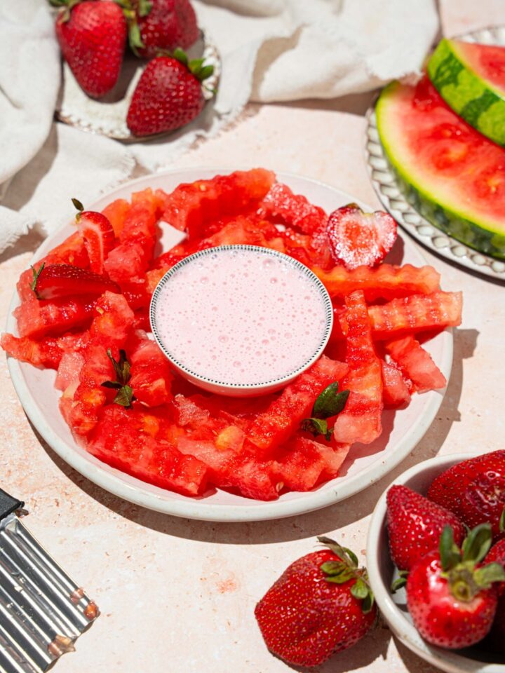 A white plate with watermelon fries and a bowl of strawberry yogurt dip in the center. The plate is garnished with sliced strawberries. The plate is on a tan counter with a bowl of strawberries and a white cloth napkin.