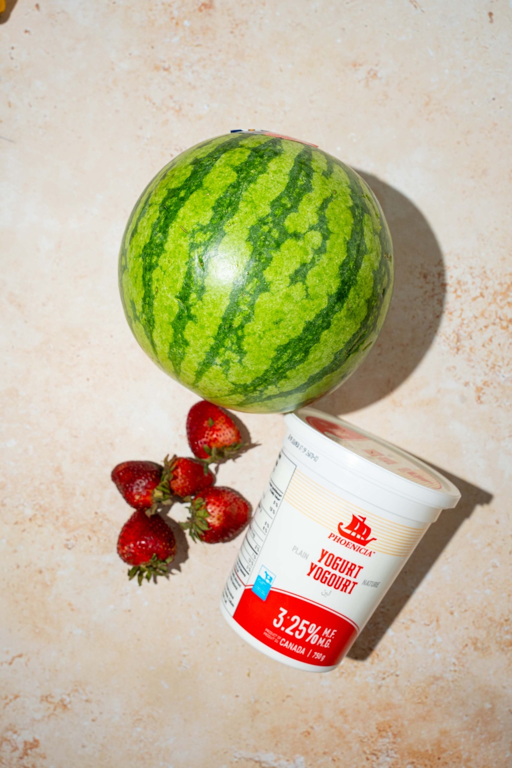 A tan counter with ingredients to make watermelon fries including a whole watermelon, strawberries, and yogurt.