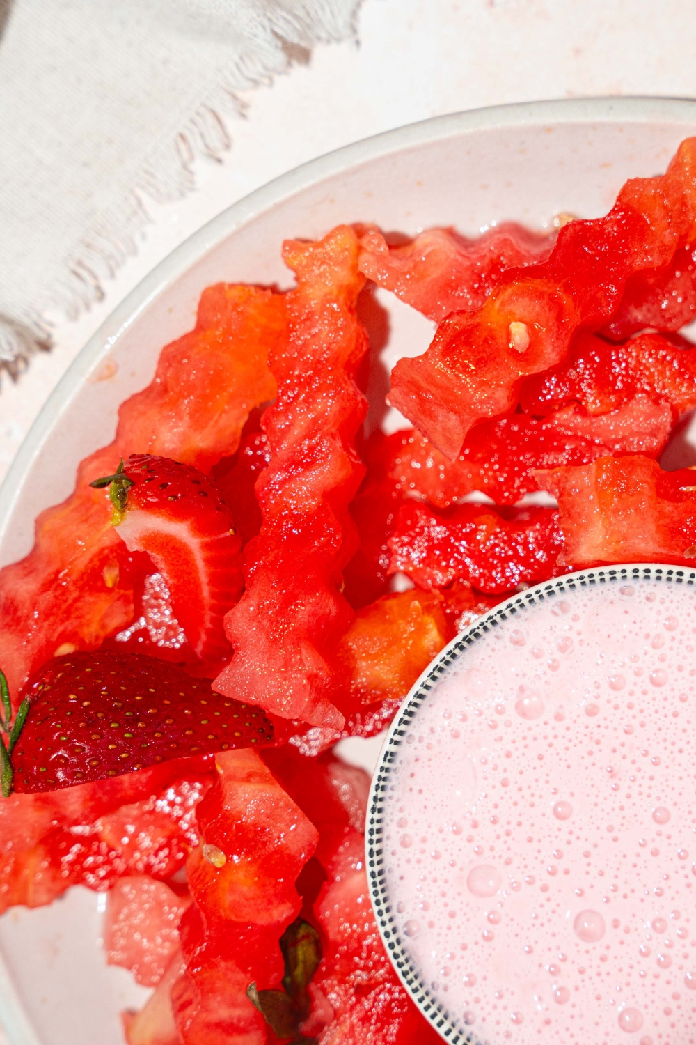 A white plate with watermelon fries and a bowl of strawberry yogurt dip in the center. The plate is on a tan counter with a white cloth napkin.