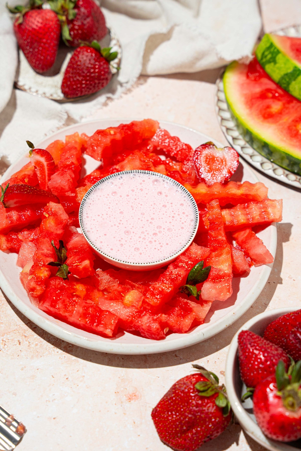 A white plate with watermelon fries and a bowl of strawberry yogurt dip in the center. The plate is garnished with sliced strawberries. The plate is on a tan counter with a bowl of strawberries and a white cloth napkin.