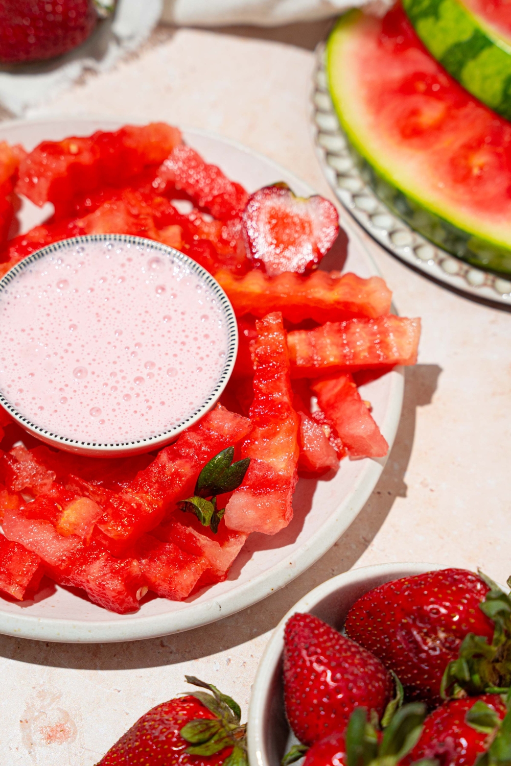 A white plate with watermelon fries and a bowl of strawberry yogurt dip in the center. The plate is garnished with sliced strawberries. The plate is on a tan counter with a bowl of strawberries and a white cloth napkin.