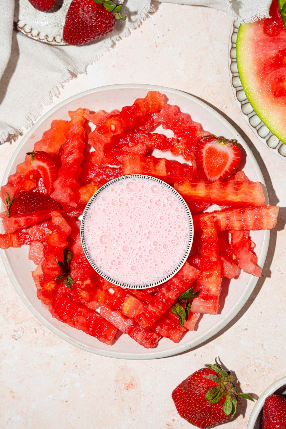 A white plate with watermelon fries and a bowl of strawberry yogurt dip in the center. The plate is garnished with sliced strawberries. The plate is on a tan counter with a bowl of strawberries and a white cloth napkin.